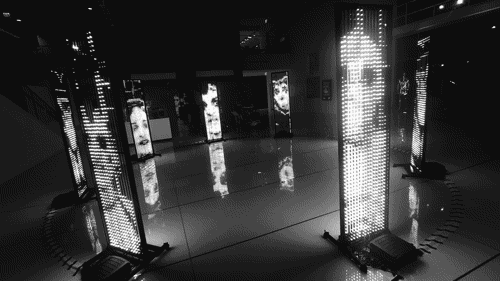 8 free-standing tall video panels form a circle in an open space, showing low resolutin images of faces and text