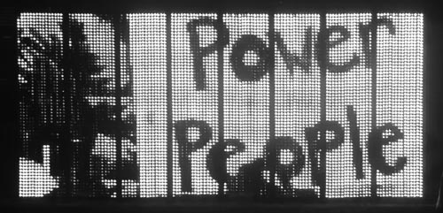 protest images animate across a two-sided free-standing large LED screen on a city river bank with people jogging and walking by
